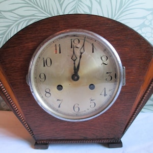 May include: A vintage wooden mantel clock with a round clock face. The clock has a dark brown wooden frame with a fan-shaped design. The clock face has black numerals and hands, and the time is approximately 12:00.
