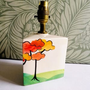May include: A triangular ceramic lamp base with a white background featuring a painted tree with orange and yellow foliage. The base has a green grassy landscape at the bottom and a brass-coloured metal light socket fixture attached to the top.