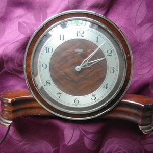 May include: A vintage wooden clock with a silver face and black hands. The clock has a wood grain pattern and is on a wooden base.