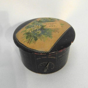 May include: A black round tin box with a floral design on the lid. The lid has the words "Forget Me Not" printed on it. The bottom of the box has a spinning pointer with the words "Spin the Pointer" printed on it.