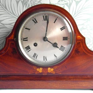 May include: A vintage wooden mantel clock with a silver face and black Roman numerals. The clock's mahogany-colored case features decorative inlays and a curved top. The clock measures approximately 25 cm wide.