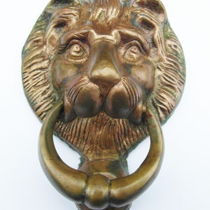 May include: A brass door knocker shaped like a lion head with a circular ring held in the lion's mouth. The metal has a weathered, aged patina with dark brown and bronze tones, showing detailed fur texture, eyes, and a prominent nose.
