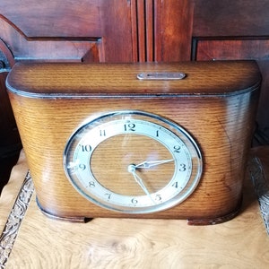 May include: A wooden mantel clock with a round, white face and black Roman numeral hour markers. The clock has a silver-coloured frame and hands.