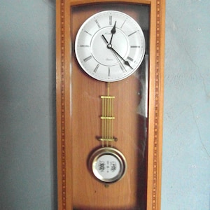 May include: A wooden wall clock with a light-coloured face, black hands, and Roman numeral hour markers. The clock has a pendulum and a decorative wooden frame. The clock is approximately 61 cm tall.