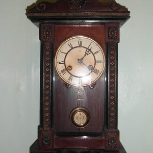 May include: An antique wall clock made of dark wood, featuring a round face with Roman numerals. The clock includes a pendulum and decorative carvings. The clock measures approximately 61 cm high and 30 cm wide.
