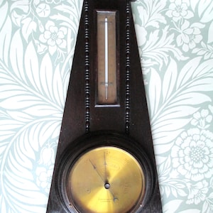 May include: A vintage wooden barometer and thermometer. The dark brown wooden frame has a triangular shape with a circular brass-coloured barometer at the bottom and a thermometer in the centre. The piece has decorative beaded accents.