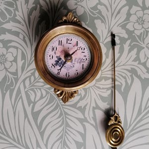 May include: A vintage wall clock with a gold-coloured frame and pendulum. The clock face has a floral design with pink and blue details. The clock is mounted on a wall with a floral pattern.
