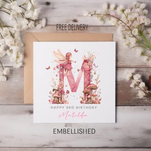 May include: A birthday card featuring a pink letter "M" adorned with a fairy, mushrooms, and flowers. The card says "Happy 3rd Birthday Matilda" and "Embellished". The background includes a neutral envelope and floral decorations.