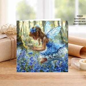 May include: A greeting card featuring an illustration of a fairy with blue wings and a blue dress kneeling in a field of blue flowers, holding a glass jar. The text on the card reads 350GSM QUALITY CARD SUPPLIED WITH KRAFT ENVELOPE.