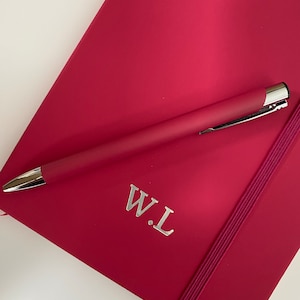 May include: A crimson red notebook with a silver elastic band closure and the initials "W.L." embossed on the front cover. A silver pen is lying on top of the notebook.