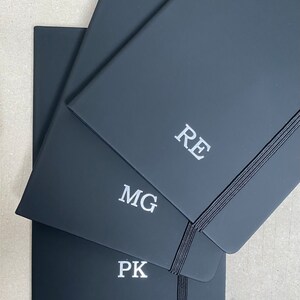 May include: Three black notebooks with elastic closures. The top notebook has the initials "RE" embossed in silver. The middle notebook has the initials "MG" embossed in silver. The bottom notebook has the initials "PK" embossed in silver.