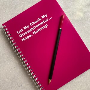 May include: A hot pink spiral-bound notebook with the text "Let Me Check My Giveashitometer... Nope, Nothing!" in white. A black pencil with a red eraser rests on the notebook.