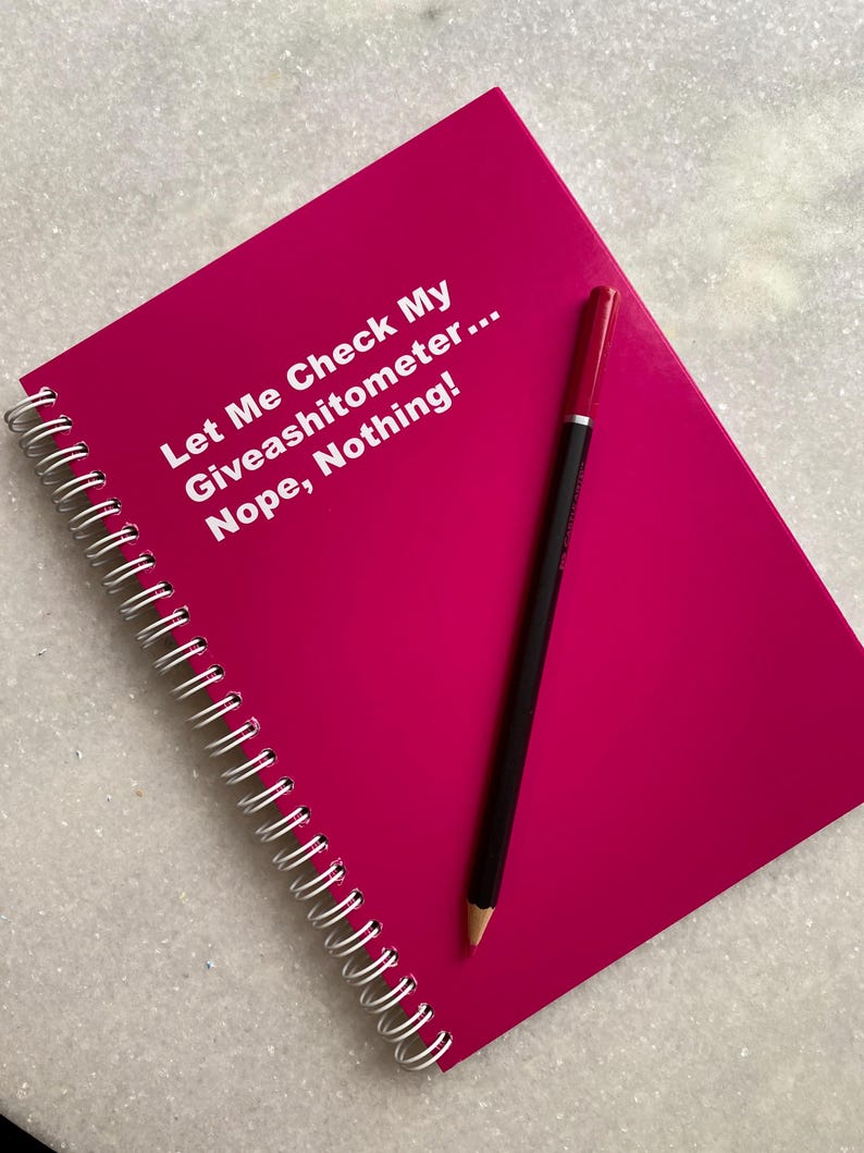 May include: A hot pink spiral-bound notebook with the text "Let Me Check My Giveashitometer... Nope, Nothing!" in white. A black pencil with a red eraser rests on the notebook.
