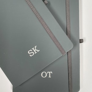 May include: Two grey hardback notebooks with elastic closures. The top notebook has the initials "SK" embossed on the front cover. The bottom notebook has the initials "OT" embossed on the front cover.