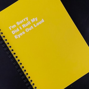 May include: A yellow spiral-bound notebook with the text "I'm Sorry Did I Roll My Eyes Out Loud" in white. The notebook is angled, set against a black background.