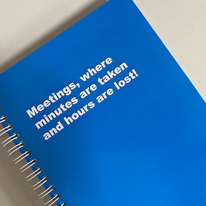 Op de afbeelding: Een blauw spiraalgebonden notitieboek met witte tekst: "Meetings, where minutes are taken and hours are lost!" Het notitieboek ligt op een lichtgrijze ondergrond.