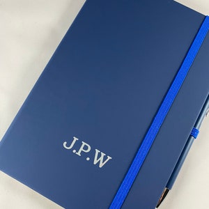 May include: A navy blue notebook with a blue elastic band closure and a silver pen. The notebook has the initials "J.P.W." embossed in silver on the front cover.