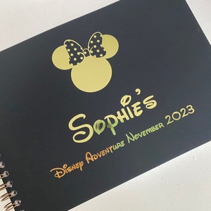 May include: Black spiral bound notebook with a gold foil Mickey Mouse head and bow, the text "Sophie's Disney Adventure November 2023" in gold foil.