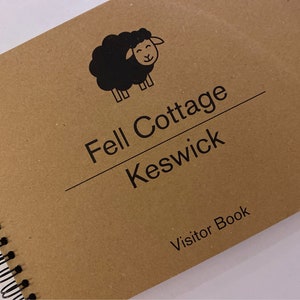 May include: A spiral-bound visitor book with a brown cover. The cover features a black illustration of a sheep and the text "Fell Cottage Keswick" and "Visitor Book" in black. The book is on a white surface.
