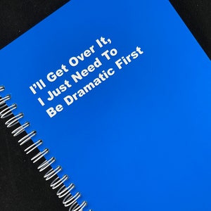 May include: A blue spiral bound notebook with the text "I'll Get Over It, I Just Need To Be Dramatic First" printed in white.