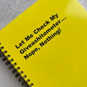 May include: A bright yellow spiral-bound notebook with black text that reads "Let Me Check My Giveashitometer... Nope, Nothing!" The notebook is open, displaying the glossy cover. The spiral binding is black.