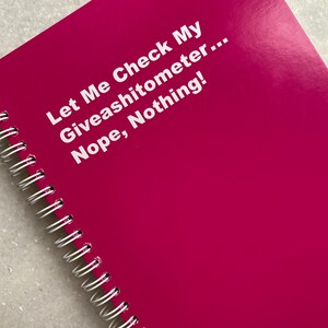 May include: A bright magenta spiral-bound notebook. The cover features the text "Let Me Check My Giveashitometer... Nope, Nothing!" in white. The notebook's spiral binding is silver.