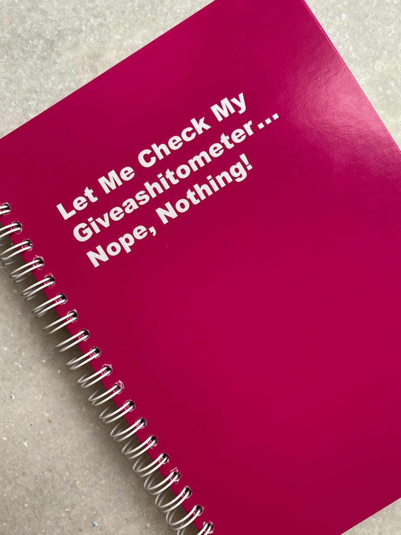 May include: A bright magenta spiral-bound notebook. The cover features the text "Let Me Check My Giveashitometer... Nope, Nothing!" in white. The notebook's spiral binding is silver.