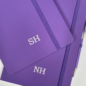 May include: Two purple notebooks with elastic closures and personalised initials, "SH" and "NH", in silver lettering.
