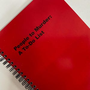 May include: A red spiral-bound notebook with the text "People to Murder: A To-Do List" printed in black. The notebook is angled, showing the spiral binding and the glossy cover.