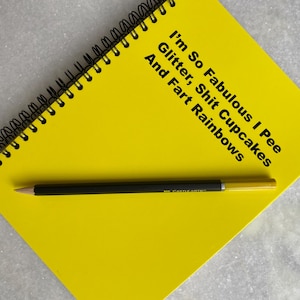 May include: A yellow spiral notebook with the text "I'm So Fabulous I Pee Glitter, Shit Cupcakes And Fart Rainbows" written on the cover. A black pencil is lying on top of the notebook.