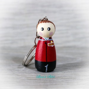 May include: A wooden doll keychain painted to look like a person wearing a red jacket with a black shirt and black trousers. The jacket has a yellow patch that says "Royal Mail" and the number "1" is painted on the trousers.