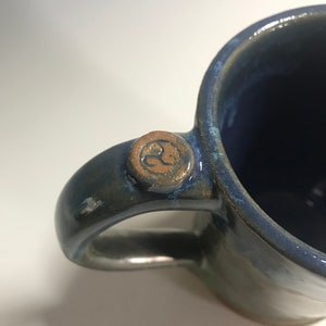 May include: A blue ceramic mug with a brown handle and a brown symbol on the handle. The mug has a glazed finish.