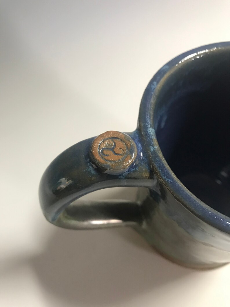 May include: A blue ceramic mug with a brown handle and a brown symbol on the handle. The mug has a glazed finish.