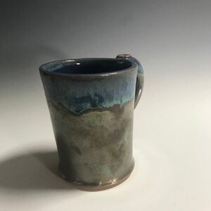 May include: A handmade ceramic mug with a blue, green, and brown glaze. The mug has a rounded handle and a wide base.