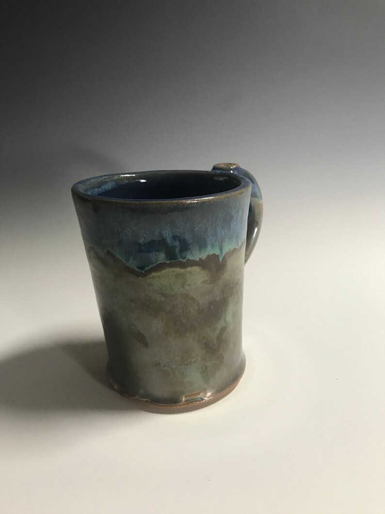 May include: A handmade ceramic mug with a blue, green, and brown glaze. The mug has a rounded handle and a wide base.