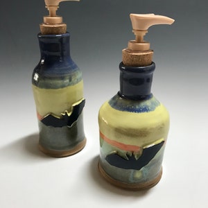 May include: Two ceramic soap dispensers with a bat design. The dispensers are hand-painted with blue, yellow, and orange stripes. The dispensers have cork stoppers and plastic pumps.