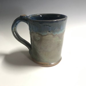 May include: A handmade ceramic mug with a blue, green, and brown glaze. The mug has a rounded handle and a wide base.