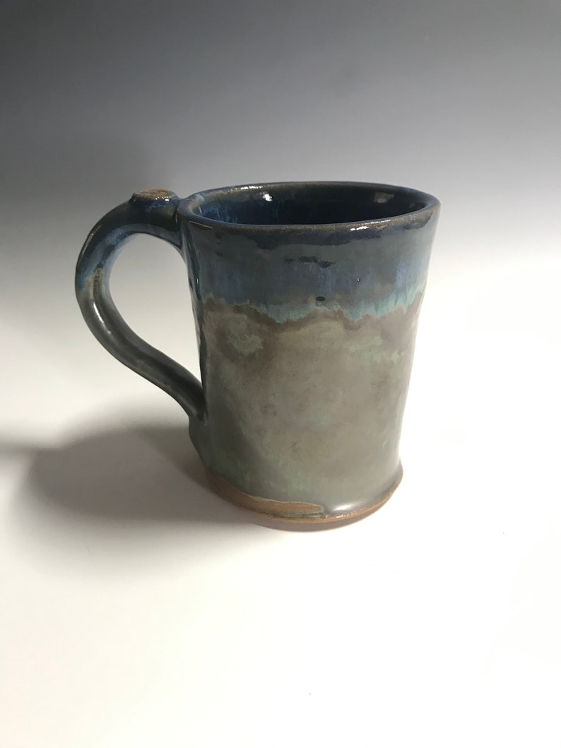 May include: A handmade ceramic mug with a blue, green, and brown glaze. The mug has a rounded handle and a wide base.