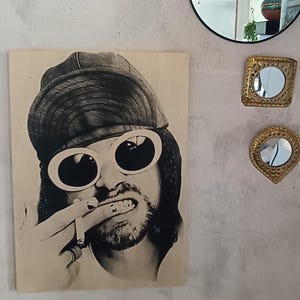 Peut inclure: Photographie en noir et blanc d'un homme portant un chapeau noir et des lunettes de soleil blanches. Il tient une cigarette dans sa bouche et a une expression sérieuse sur son visage.