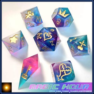 May include: A collection of seven polyhedral dice with a fantasy theme. The dice are made of clear resin with a galaxy-like effect in shades of blue, purple, and pink. Gold-colored symbols and numbers are embedded within the dice.