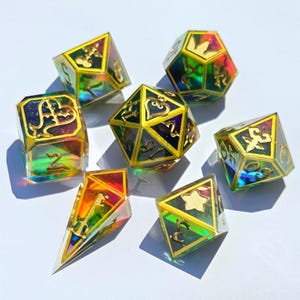 May include: A set of seven polyhedral dice featuring a translucent, iridescent rainbow finish with deep purple and blue internal swirls. Each die has sharp, gold-painted edges and gold-filled symbols, including hearts, stars, and crowns.
