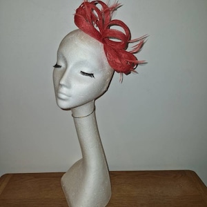 May include: A coral coloured fabric flower hair accessory with feathers. The flower is in a bow shape and is on a white mannequin head.