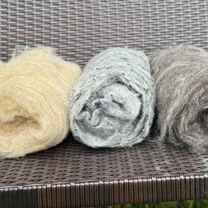Può includere: Cinque rotoli di lana di diversi colori, tra cui bianco, beige, grigio, grigio scuro e nero. La lana è disposta su una superficie in vimini intrecciato.
