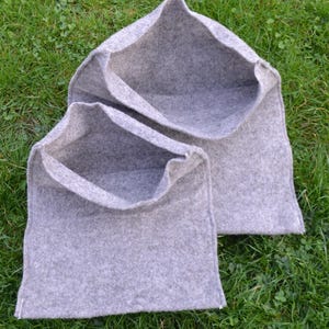 May include: Two grey felt storage bags with open tops, set on green grass. The bags are rectangular with a pointed top and white stitching along the bottom edges. The felt has a soft, textured appearance.