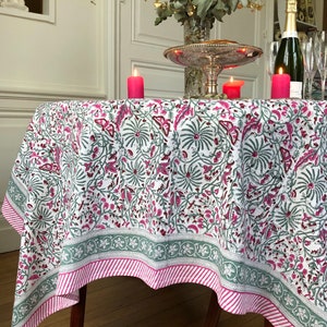 May include: A white tablecloth with a pink and green floral pattern. The tablecloth has a green and pink striped border. The tablecloth is laid on a round table with a wooden base. There are three pink candles on the table.