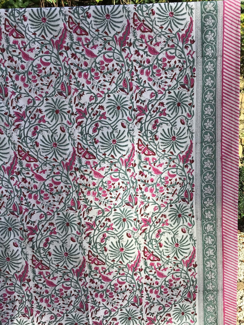 May include: White fabric with a pink and green floral pattern. The fabric has a border with a repeating floral design.