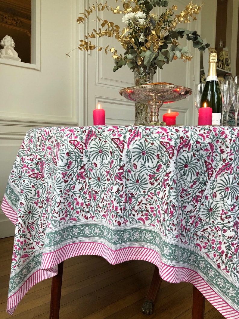 May include: A white tablecloth with a pink and green floral pattern. The tablecloth has a green and pink striped border. The tablecloth is laid on a round table with a wooden base. There are three pink candles on the table.
