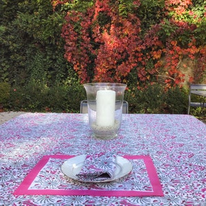 May include: A white tablecloth with a pink and gray floral pattern. The tablecloth is set with a white plate, a pink napkin, and a glass candle holder with a white candle.