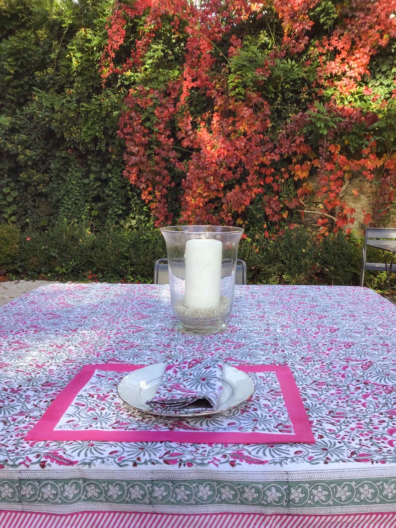 May include: A white tablecloth with a pink and gray floral pattern. The tablecloth is set with a white plate, a pink napkin, and a glass candle holder with a white candle.