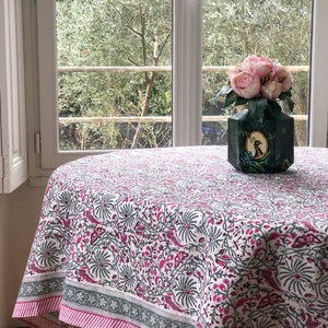 May include: A white tablecloth with a pink and gray floral pattern. The tablecloth has a pink and gray border. A vase with pink flowers sits on the tablecloth.
